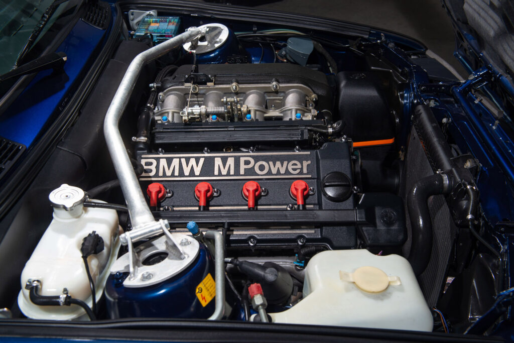BMW M3 E30 Sport Evolution in Mauritiusblau (AC79200) — Motorraum mit vier roten Zündkerzensteckern, Ansaugtrakt und BMW-M-Power-Ventildeckel