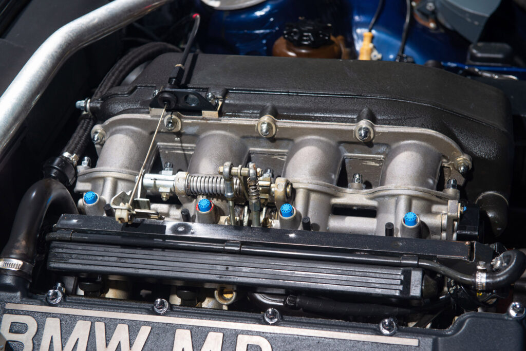 BMW M3 E30 Sport Evolution in Mauritiusblau (AC79200) — Nahaufnahme des Ansaugtrakts mit Einzeldrosselklappen über dem M-Power-Ventildeckel