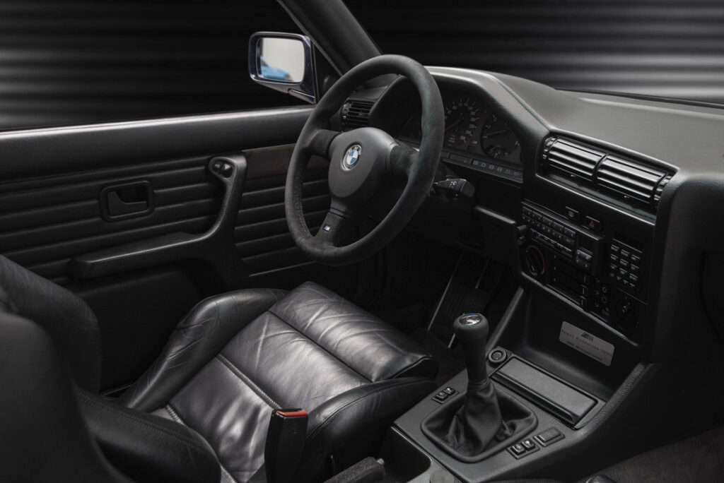 BMW M3 E30 Sport Evolution in Mauritiusblau (AC79200) — Cockpit mit Lenkrad, Armaturenbrett und Schalthebel von der Beifahrerseite